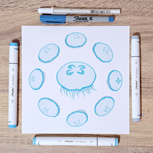 Staged photo of the "Jellyfish Lunar Cycle" illustration on a wooden surface. There are various pens and markers surrounding the paper. The jellyfish are light blue with white details.