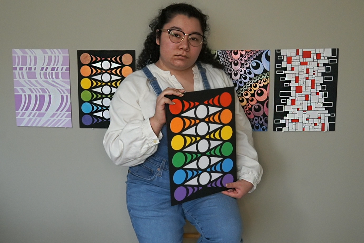 Staged photo of Peacein sitting on a stool holding the "Eye Chevron" painting in front of a wall with several abstract paintings hung up on an off white wall